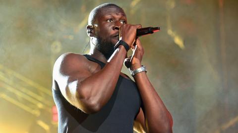 A man in a black vest performing at a live music concert. He holds a microphone up to his mouth.