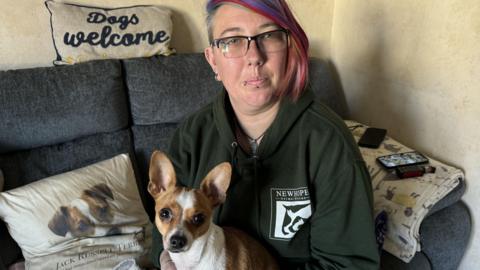 Emily sits in a green hoodie wearing glasses and holding Tilly, a small Jack Russell cross Chihuahua.