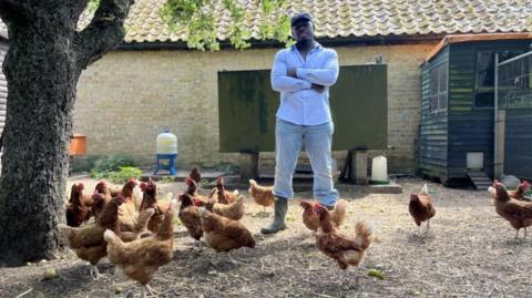 Fekky, wearing a navy blue cap, white shirt, jeans and wellington boots, stands with his arms folded looking at camera. He is surrounded by chickens on his farm.