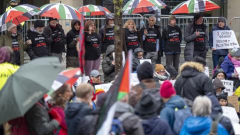 Crowd gathers in front of a line of people under umbrellas wearing Palestine Action T-shirts. A man at the end of the line holding a white poster which says "I OPPOSE GENOCIDE. I SUPPORT PALESTINE ACTION"