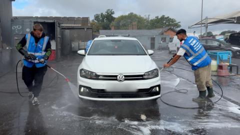 Image shows a white Volkswagen Polo in the centre. It is being washed. Either side of the car is a worker holding a jet wash. They are washing off the tyres. The worker on the left is wearing a black tracksuit and a blue high-vis. The worker on the right is wearing beige trousers and a white top. He is also wearing a blue high-vis. Behind the car are two grey buildings.