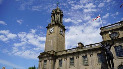 The exterior of South Shields Town Hall with the clock tower in the centre of the image. It is an ornate, stone-built structure. An England flag is flying from another part of the building. The clockfaces sit on all four sides of the tower (only two are seen in the image) about halfway up. The tower itself has flat sides made of large stone until just above the clockfaces where it gives way to far more ornate masonry including statues and columns.