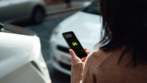 A woman with long hair holds a mobile phone showing a generic taxi app, in front of two silver cars 