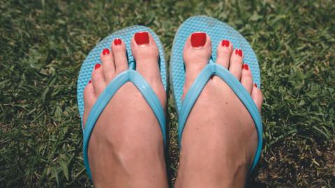 A pair of feet with painted toenails wearing blue flip-flops is seen against green grass.
