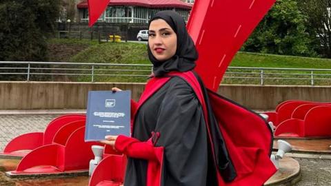 Llun o Zaina Aljumma wedi graddio ac yn sefyll o flaen draig goch.