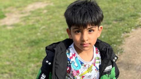 A young boy called Saleh looks directly to camera. He has dark hair and is wearing a black and green coat over a multi coloured t-shirt. He is stood on a concrete path and to the left of the background is grass. 