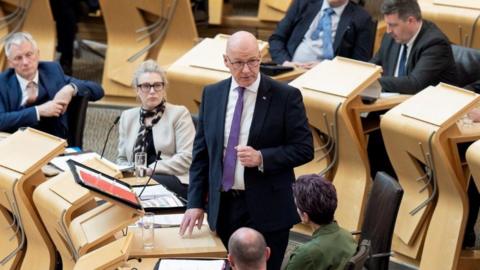 John Swinney stands in the Holyrood chamber surrounded by sitting MSPs