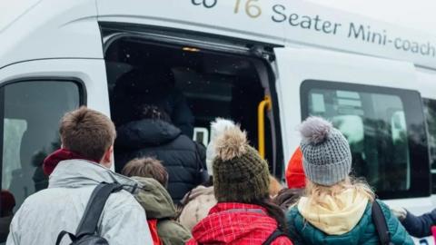 A white minibus with "16 Seater Mini-coach" written above the door. A sliding door is open, with a yellow handrail on one side. Children in winter clothing are boarding the bus.