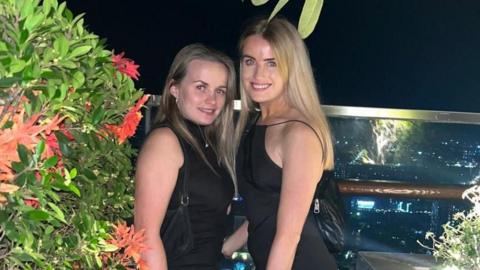 Bethany Clarke and Simone White while travelling. The friends, who both have long blonde hair, wear matching black dresses and are pictured on a terrace at night time. They smile at the camera, surrounded by foliage and a view of a city behind them. 