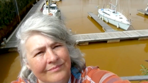 Marianne Birkby is taking a selfie in front of the orange water at Whitehaven Harbour. The harbour has a wooden pontoon, and two boats docked. Birkby has mid-length grey hair and is wearing an orange jacket.