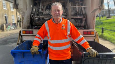 Chris Lively looking into camera. He has short hair and is wearing an orange council jumper, and yellow gloves. He is standing in between two bins, which he is holding, and in front of the back of a bin lorry.