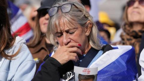 A woman with blonde hair, draped in a blue and white Israeli flag, closes her eyes and bows her head, with one hand held over her mouth. In the other, she holds a flyer displaying a picture of a male Israeli hostage, along with his name - David Cunio - and age - 34 - printed in red, capital letters, in both English and Israeli. Other attendees are pictured out of focus in the background.