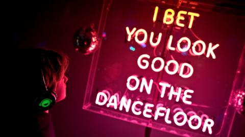 A pink lit up sign which reads 'I bet you look good on the dancefloor' in a clear, square casing. There is a small disco ball to the left and a young boy with short hair and lit up over-the-ear headphones looking at the sign. He is partly in shadow. The photo is slightly on an angle and the sign is lighting up the room in a pink light.