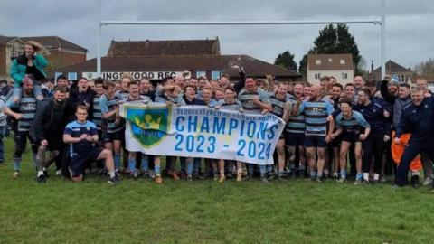 Kingwood RFC men in their sky blue and dark brown hooped rugby shirts. They are hold a banner proclaiming them 2023/2024 Gloucestershire Tribute South champions. All are smiling and several are punching the air in celebration.
