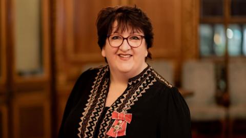 A smiling woman wearing glasses stands in a formal wood‑panelled room, dressed in a black embroidered outfit with a red‑and‑gold medal pinned to her chest, suggesting they have just received an honour or award.