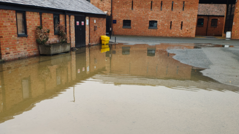 A flooded road with a small red brick building on the left with black doors and windows and plants at the front. There's another larger red brick building behind it. 