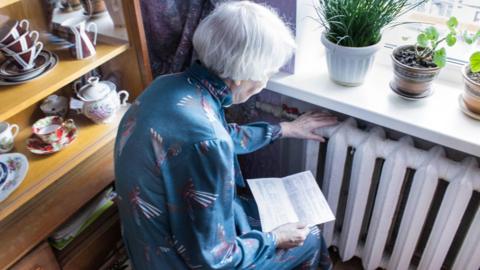 A stock image of an elderly woman holding an energy bill while sat close to a radiator.