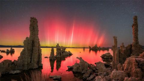 A bright pink and yellow aurorae shine over a lake with rocks. The sky behind is dark and covered in lots of tiny stars.
