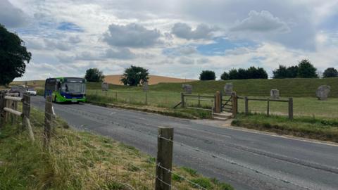 The road running through Avebury with traffic, including a bus, approaching. Behind the road can be seen a field with part of the large stone circle visible, and behind those stones is a grassy embankment with trees in the distance. 