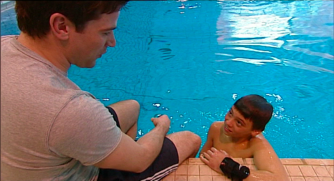 Gethin Jones on the left hand side, sitting on the edge of the side of a swimming pool.  Tom Daley is in the pool on the right hand side, leaning on the edge looking up at Gethin.