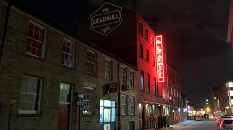 A four-storey red brick building with a black sign running vertically along the wall with the words 'The Leadmill' in red, neon writing. 