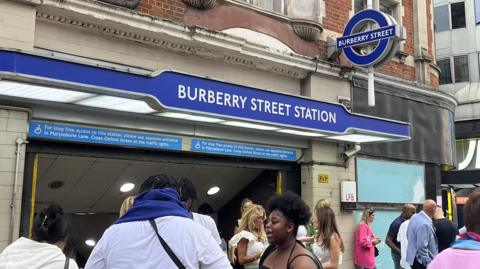 People walking in and out of an underground station with a blue sign above the entrance. The name written on the sign in white capital letters is "Burberry Street Station". Sticking out from the wall of the building above is a blue circular sign with "Burberry Street" written in white capital letters on a strip across the middle.