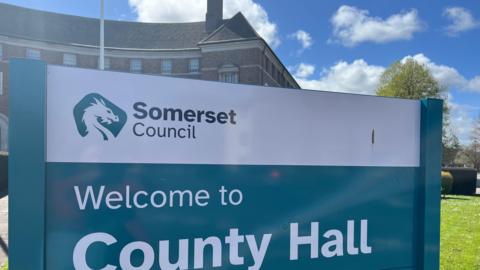 The sign for Somerset Council's county hall. It is blue and white. In the background is part of the building and some grass.