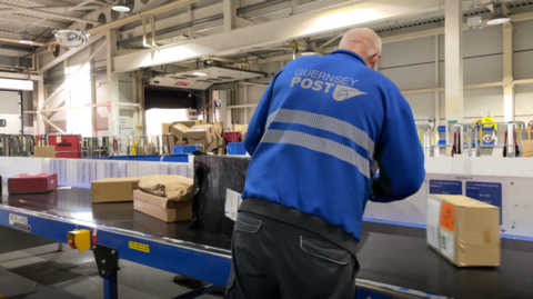 Guernsey Post staff member wearing a branded vest standing at a conveyor belt looking away from the camera