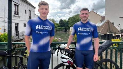 James Howell on the left with riding partner Max Edginton. They are each wearing blue and white cycling clothes and are stood in front of their bikes