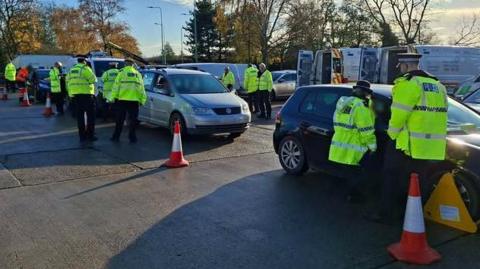 A number of police officers, and other people in high-vis jackets, at a checkpoint. There are cars lined up. The one at the front has an orange wheel-clamp on it. There are red traffic cones on the ground, and lots of vehicles parked up. 