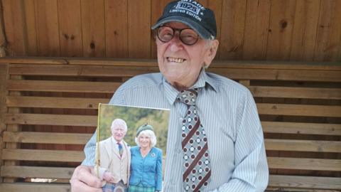 Bill sits in a wooden hut and is wearing a blue and white striped shirt, and grey, white and maroon tie with stripes and dots on it, and dark grey baseball cap with a logo and some writing on it, one bit of which says 1925. He is holding up a photo of the King with his wife, Queen Camilla towards the camera.