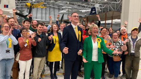 A crowd of people wearing Liberal Democrat rosettes cheer and celebrate. 