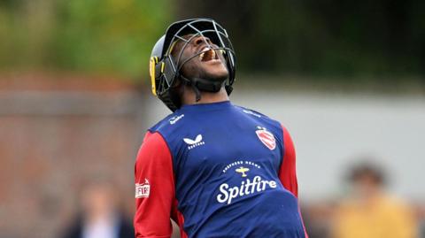 A mid shot of Daniel Bell-Drummond. He is wearing a protective cricket helmet, and a blue shirt with red sleeves. He is roaring with joy after scoring for Kent.