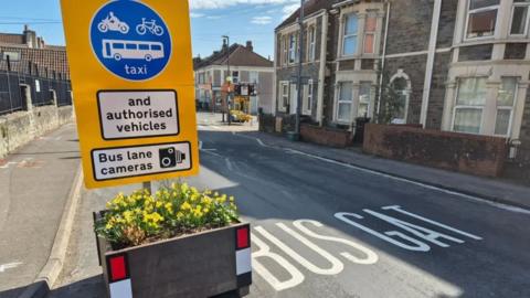 A road is partially blocked by a planter with a sign above it which indicates only buses, taxis, motorbikes and bicycles can pass. The words 'BUS GATE' are written on the road itself. It is a residential street with terraced houses.