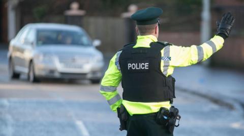 A generic photograph of a policeman dressed in high visibility yellow jacket and black vest with the word police written in silver capital letters on the back. He is wearing black gloves and raising his arm to stop a silver car which is approaching on the road.