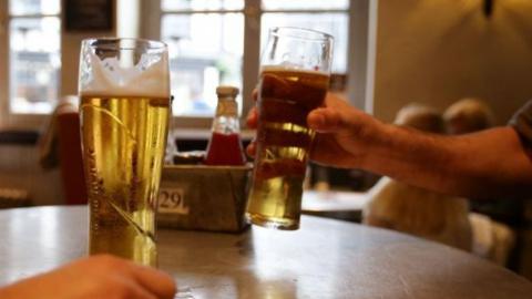 A pub table with two pints of cider. One man goes to lift his drink, you can only see his arm. A tub with a bottle of ketchup also rests on the table, with the number 29 attached to it.