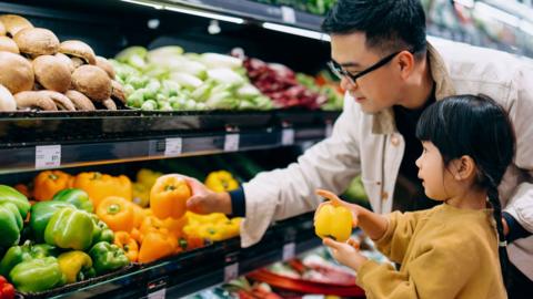 Man with black hair and glasses wearing a white jacket is leaning over the vegetable aisle in the supermarket holding a yellow pepper. There is a young girl with him who is holding a yellow pepper. They are choosing vegetables together. They look as if they are father and daughter. The daughter has black hair tied in a plait and is wearing a yellow top.