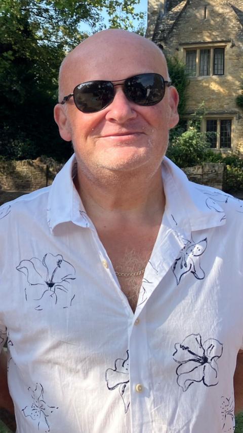 A man in a white shirt and sunglasses smiles to the camera.
