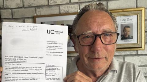 A head and shoulders image of a man wearing a grey polo shirt and glasses. He is holding a letter from Universal Credit while looking into the camera.