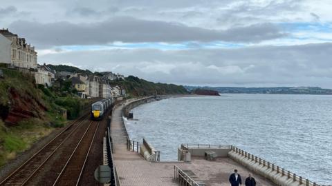 Sea wall in Dawlish. There are train tracks and a train on the left, and a pathway on the right. To the right of the trainline are houses and a cliff face.