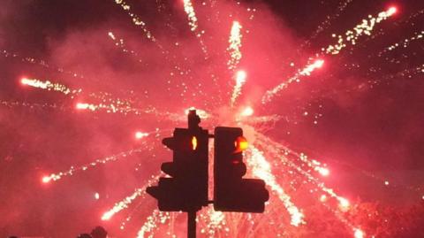 A red firework explodes behind a set of traffic lights in the night sky.