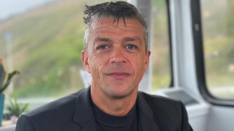 Lorne Castle smiles at the camera. He has gelled, grey hair which is short at the sides. He wears a dark jacket over a black, collarless top