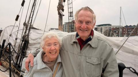 Peter has his arm around Pat and they both look incredibly happy. A boat is behind them and you can see other buildings on the other side of the dock.