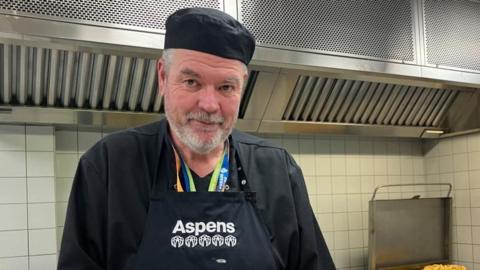 Ian Williams, wearing a black top and apron, standing in a kitchen in front of a pizza and some croissants