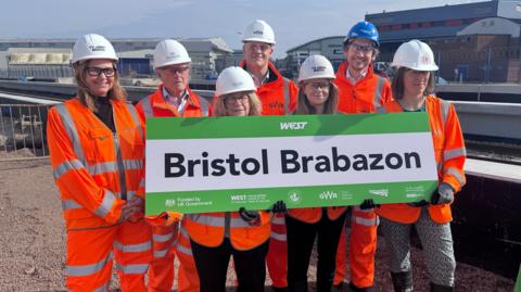 Seven people dressed in orange high-visibility clothing and hard hats, holding a giant sign which reads Bristol Brabazon. Behind them is a railway line and industrial buildings
