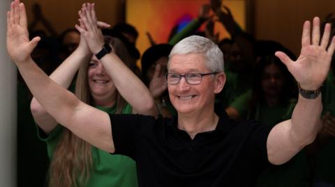 Apple CEO Tim Cook, in a black polo and smiling wide as he waves with both hands in the air at a crowd outside of a new Apple retail store. 