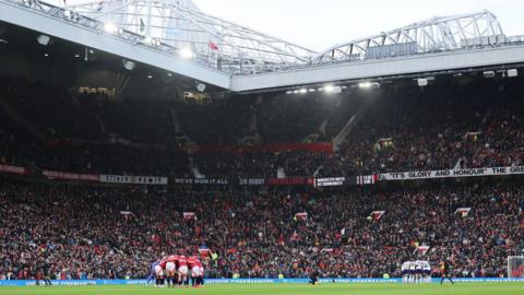 Manchester United players in a huddle in the middle of the pitch with a full Old Trafford in the background