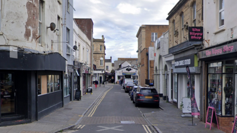 A Google maps street view of West Street in Weston-super-Mare. There are businesses either side of the street and cars parked on one side of the road.