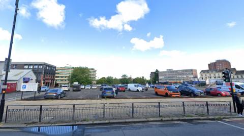 A tarmac outdoor car park in a city centre with several cars and vans sitting on it. There is a metal fence at the front with several tower blocks in the distance.