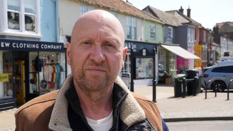 A head and shoulders image of a man wearing a light brown jacket over a black jumper and white T-shirt. He is sitting in a town centre and looking into the camera. 